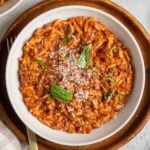 White bowl of creamy ground beef and orzo with fresh basil and freshly grated parmesan cheese on top on a dark wood plate with a gold fork and a beige and white checkered tea towel.