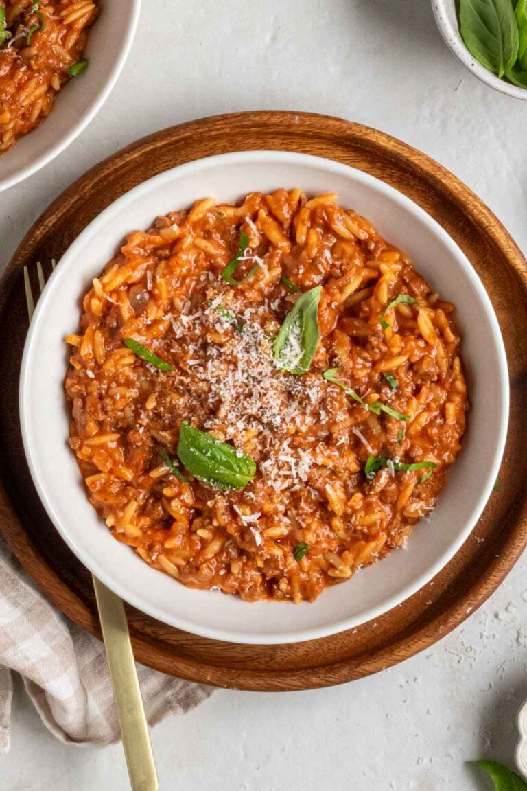 White bowl of creamy ground beef and orzo with fresh basil and freshly grated parmesan cheese on top on a dark wood plate with a gold fork and a beige and white checkered tea towel.