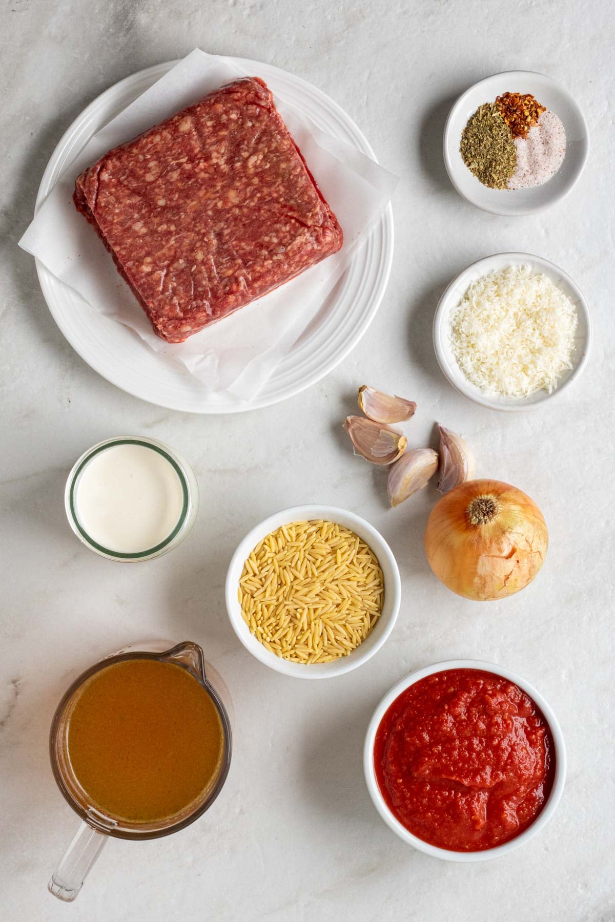 Plate of lean ground beef, bowls of spices, bowl of parmesan cheese, jar of heavy cream, bowl of orzo, onion and garlic, jug of beef broth, and a bowl of crushed tomatoes.