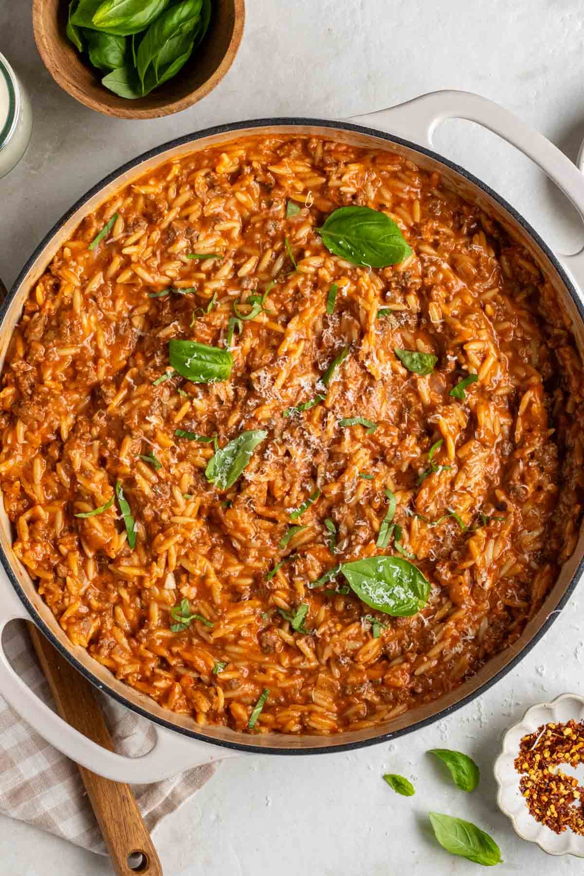 Pot with creamy ground beef and orzo pasta with freshly grated parmesan cheese and fresh basil on top with a wooden serving spoon and small bowl of red pepper flakes on the side.