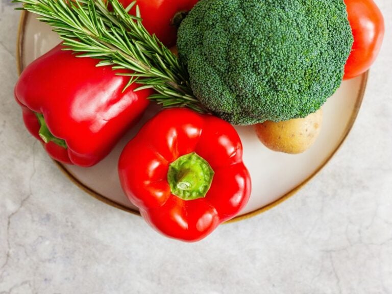 White plate with a red bell peppers, rosemary, broccoli, potatoes, and tomatoes on a counter top.