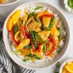 Bowl of healthy orange chicken stir fry on a bed of white rice with sesame seeds, sliced green onion, and orange slices on the side with a blue and white stripped tea towel and a fork.
