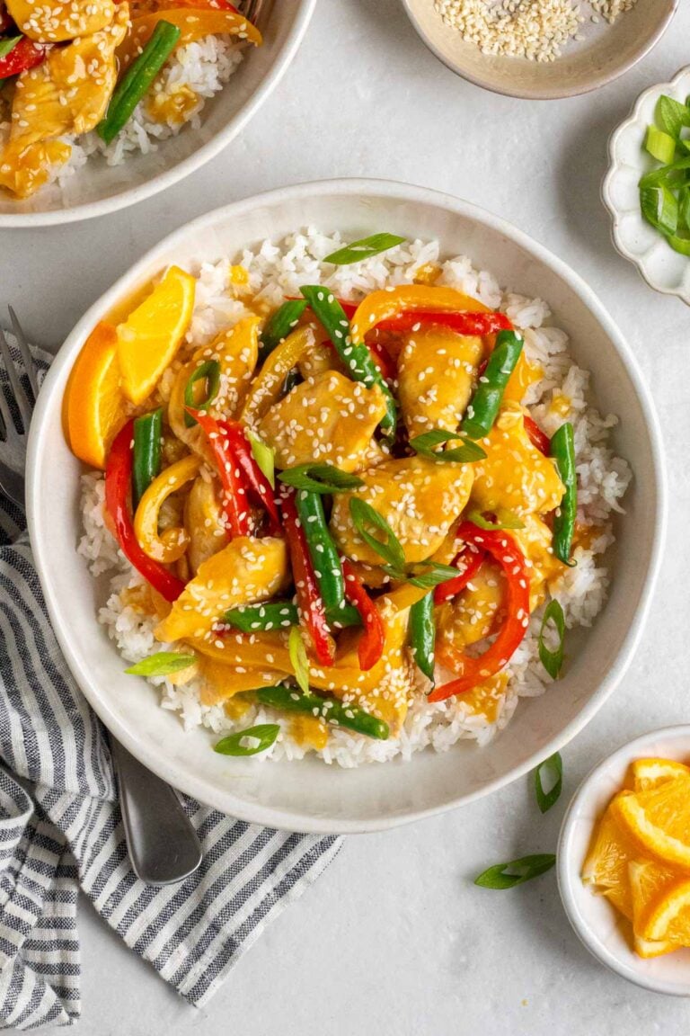 Bowl of healthy orange chicken stir fry on a bed of white rice with sesame seeds, sliced green onion, and orange slices on the side with a blue and white stripped tea towel and a fork.