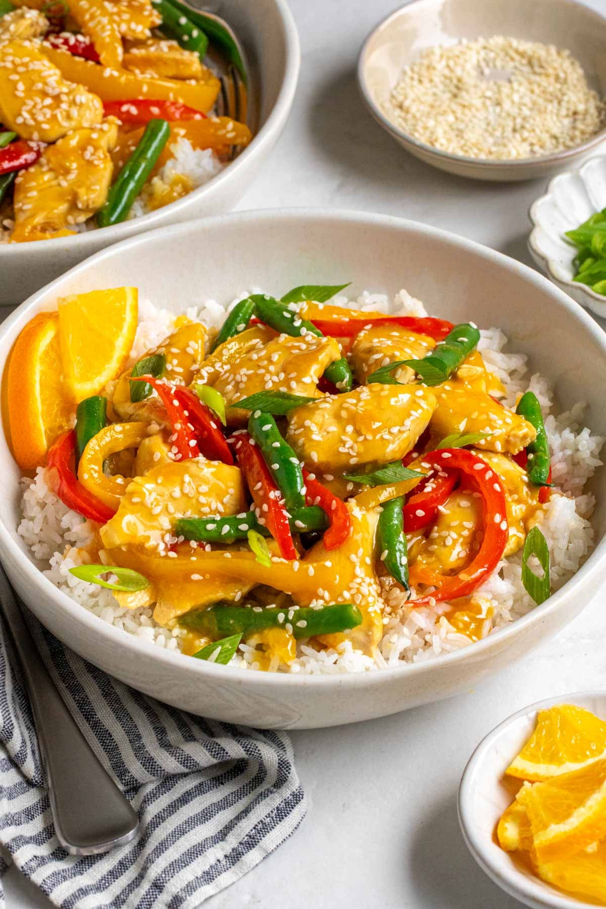 Close up of a bowl of orange chicken stir fry with white rice and sesame seeds and sliced green onions on top.