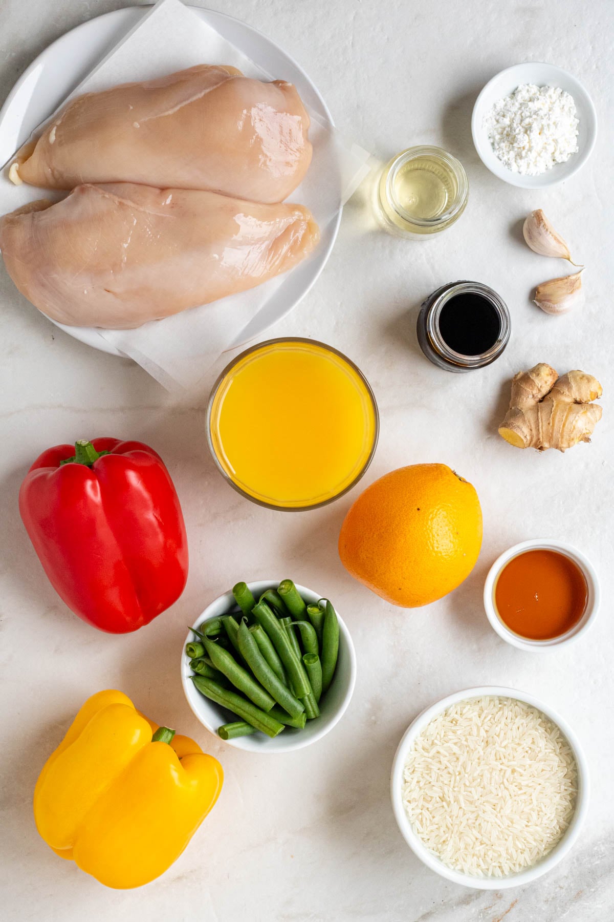 Plate of chicken breasts, bowl of cornstarch, jars or soy sauce and rice vinegar, garlic, ginger, glass of orange juice, orange, bowl of honey, bell peppers, green beans, and bowl of white rice on a white background.
