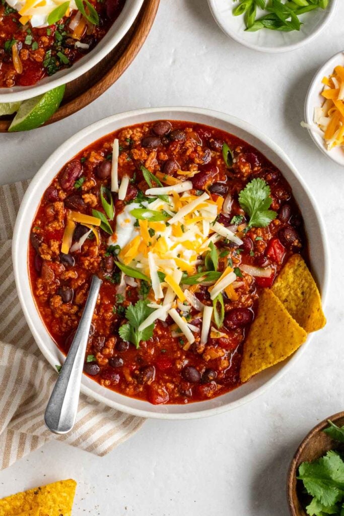 Two bowls of slow cooker turkey chili with sour cream, shredded cheese, fresh cilantro, and sliced green onion on top with tortilla chips on the side.