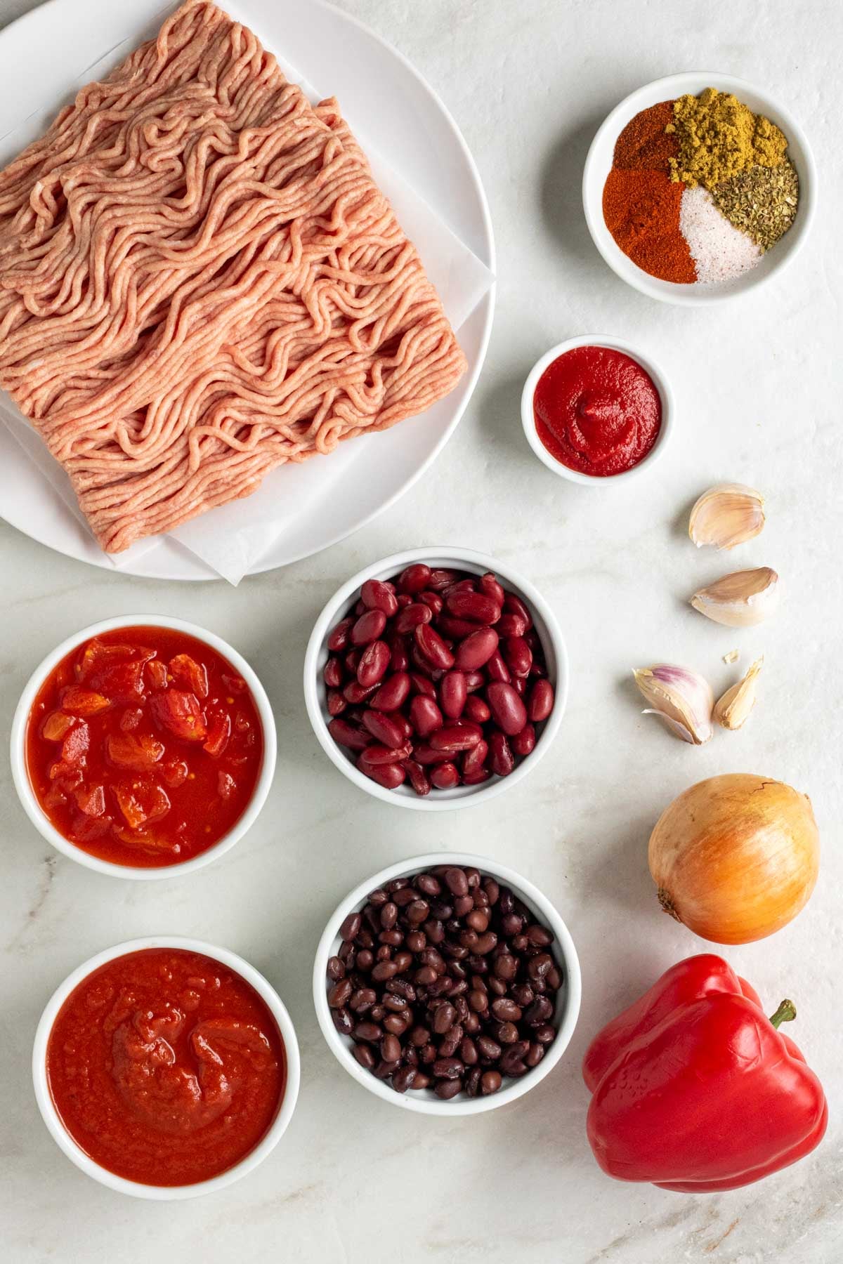 Ground turkey on a white plate, bowl of spices, bowl of tomato paste, bowl of kidney beans, bowl of black beans, bowl of diced tomatoes, bowl of crushed tomatoes, garlic cloves, onion, and red bell pepper on a white background.