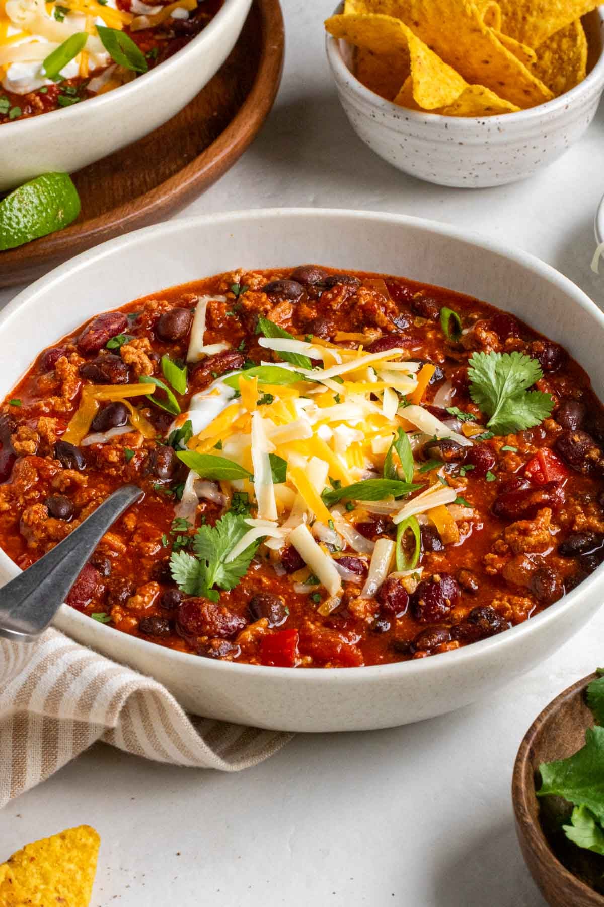 Close up of a bowl of crockpot turkey chili with a silver spoon on a beige and white striped tea towel with sour cream, cilantro, cheddar cheese, and green onions on top.