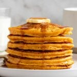 Stack of sweet potato pancakes with a knob of butter and maple syrup on top on a white plate with a glass of milk and cup of coffee in the background.