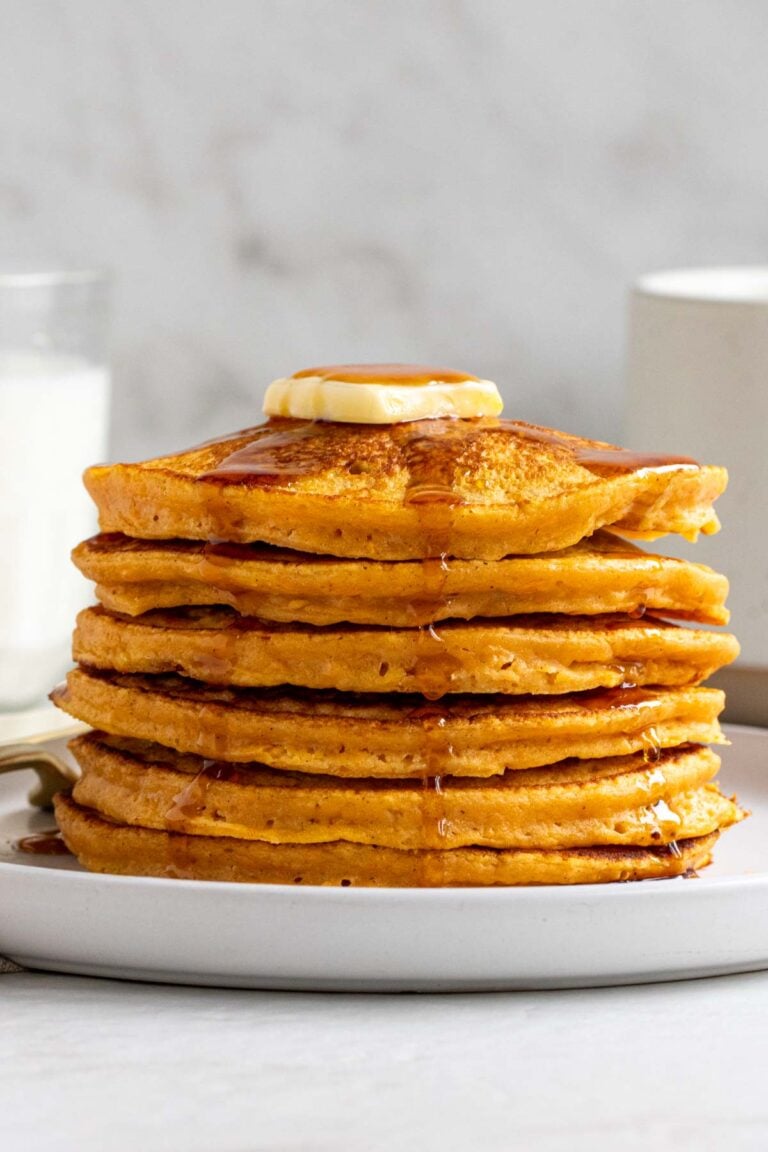 Stack of sweet potato pancakes with a knob of butter and maple syrup on top on a white plate with a glass of milk and cup of coffee in the background.