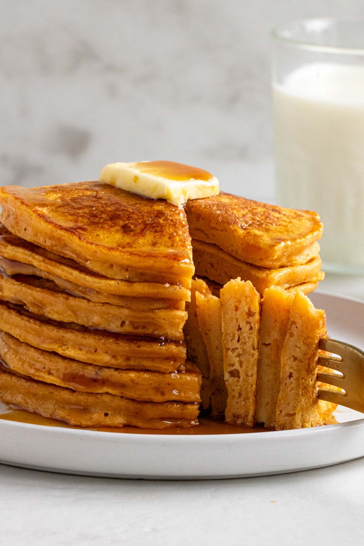 Close up of a pile of sweet potato pancakes with a slice cut out on a gold fork with butter and maple syrup on top and a glass of milk in the background.