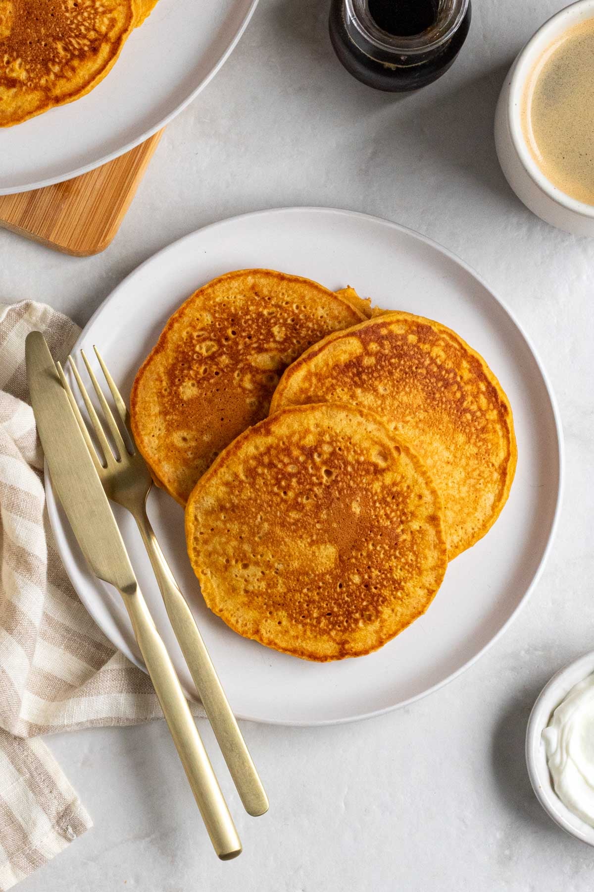 Plate with three sweet potato pancakes with a gold fork and knife and a striped beige tea towel with a jar of maple syrup, cup of coffee, and bowl of Greek yogurt on the side.