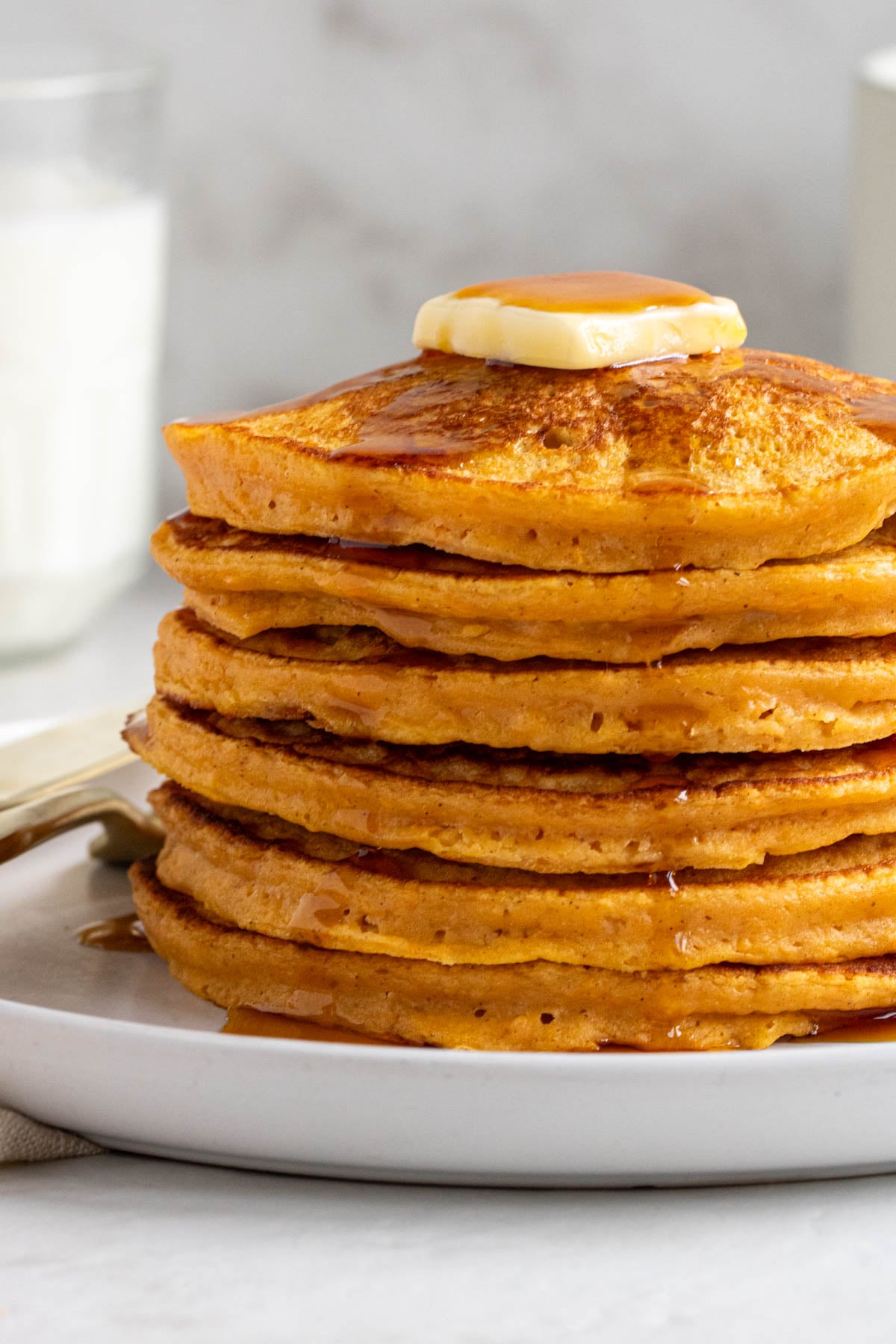 Close up of a stack of sweet potato pancakes with maple syrup and butter on top with a glass of milk in the background.