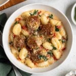 Bowl of Tuscan chicken meatballs with gnocchi on a green tea towel with a silver fork with a small bowls of sun-dried tomatoes and salt and pepper on the side.