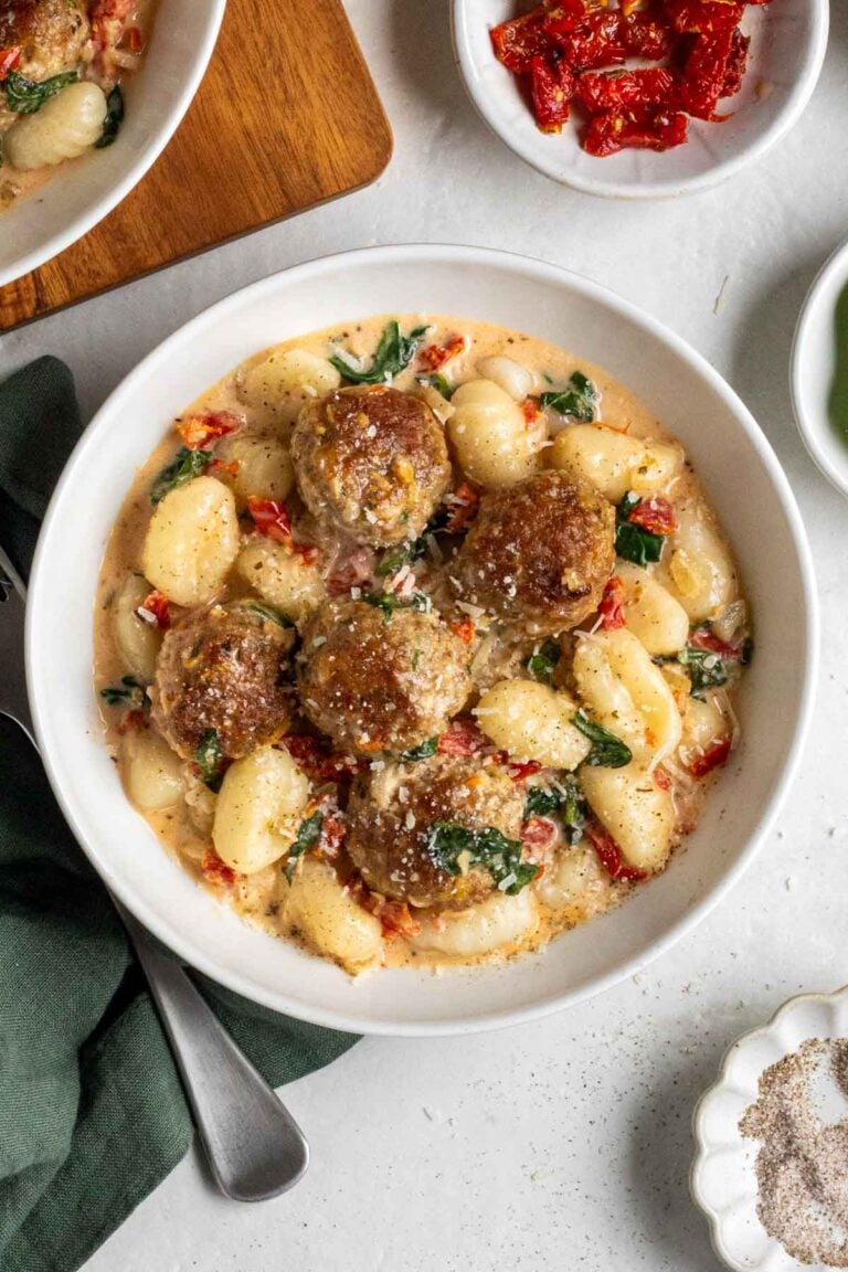 Bowl of Tuscan chicken meatballs with gnocchi on a green tea towel with a silver fork with a small bowls of sun-dried tomatoes and salt and pepper on the side.
