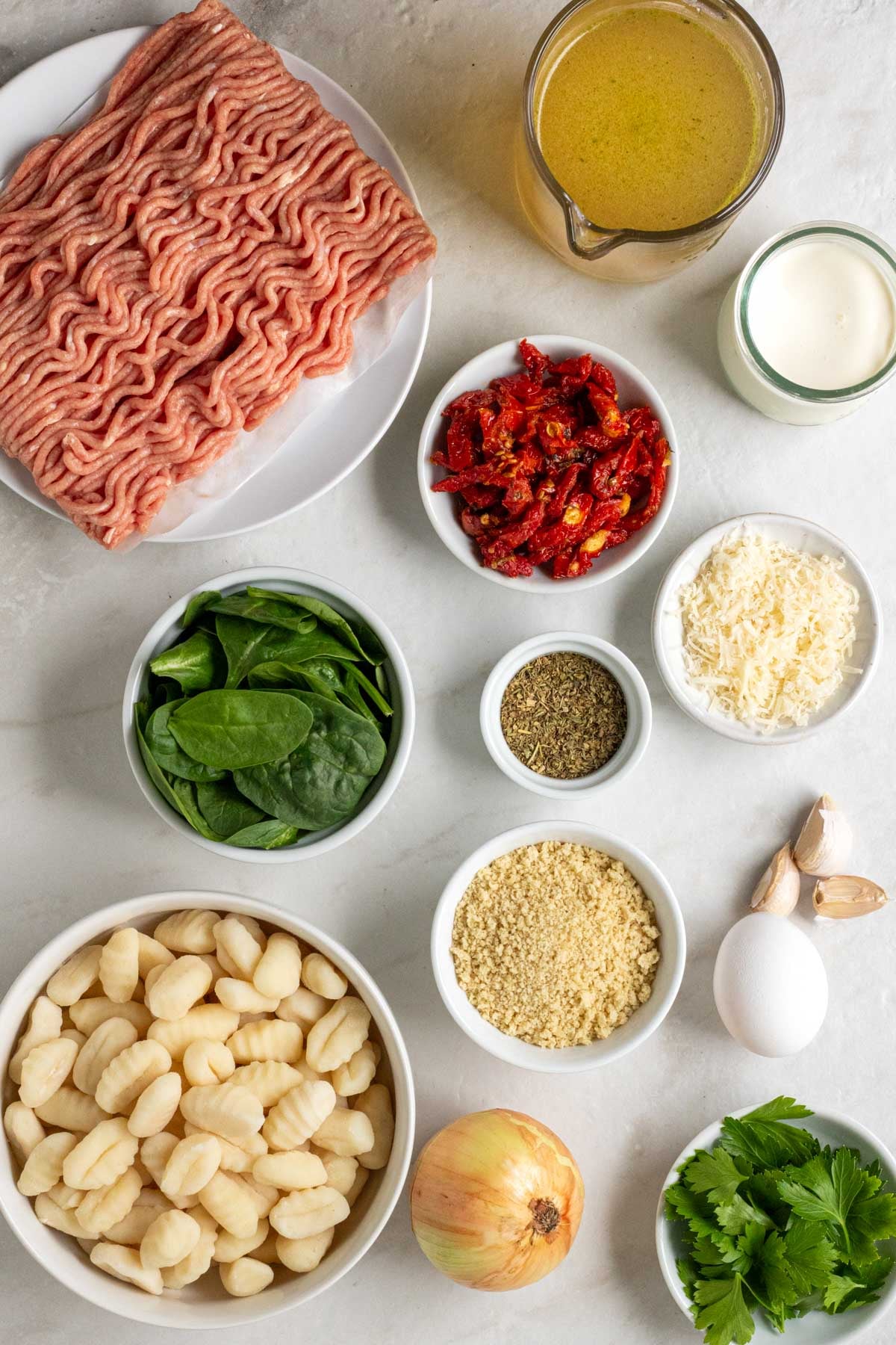 Plate of ground chicken, jug of chicken broth, jar of heavy cream, with bowls of sun-dried tomatoes, parmesan cheese, Italian seasoning, fresh spinach, breadcrumbs, and gnocchi with an garlic cloves, an egg, onion, and bowl of fresh parsley on a white background.