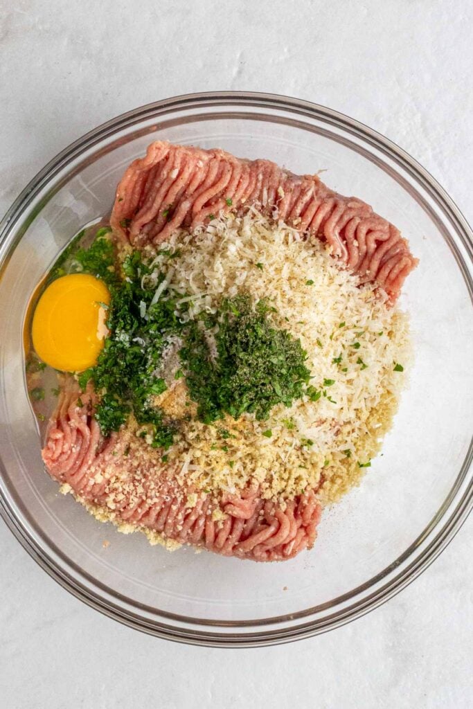 Glass bowl of ground chicken, breadcrumbs, parmesan cheese, parsley, salt, pepper, and an egg on a white background.