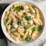 Bowl of chicken and broccoli pasta on a green tea towel with a silver fork and a small bowl of parmesan cheese and chopped parsley on the side.