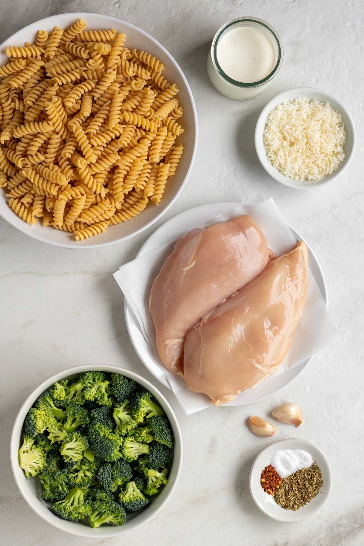 Bowl of pasta, jar of cream, bowl of parmesan, plate of chicken breasts, bowl of broccoli florets, garlic cloves, and a bowl of spices on a white background.