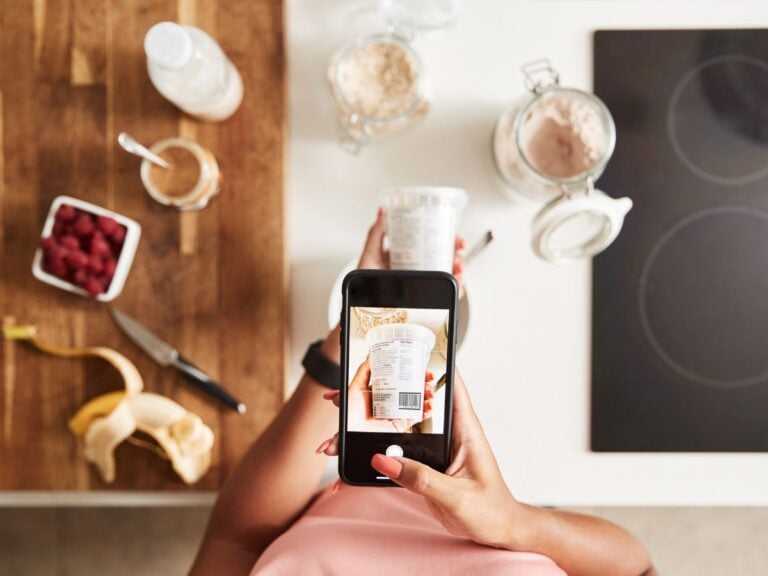 Overhead image of a woman holding an iPhone while scanning a tub of yogurt with raspberries, peanut butter, a banana, and oatmeal on a kitchen counter.